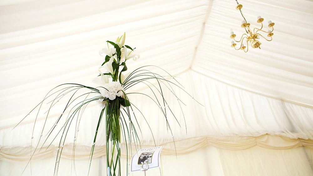 Cream wedding marquee lining with dainty chandelier to ceiling and thin flower vase with lilies and draped tendrils