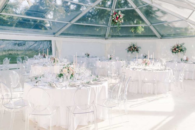 Garden Wedding Marquee with clear roof and see through chairs sat around white dressed tables with floral arrangements