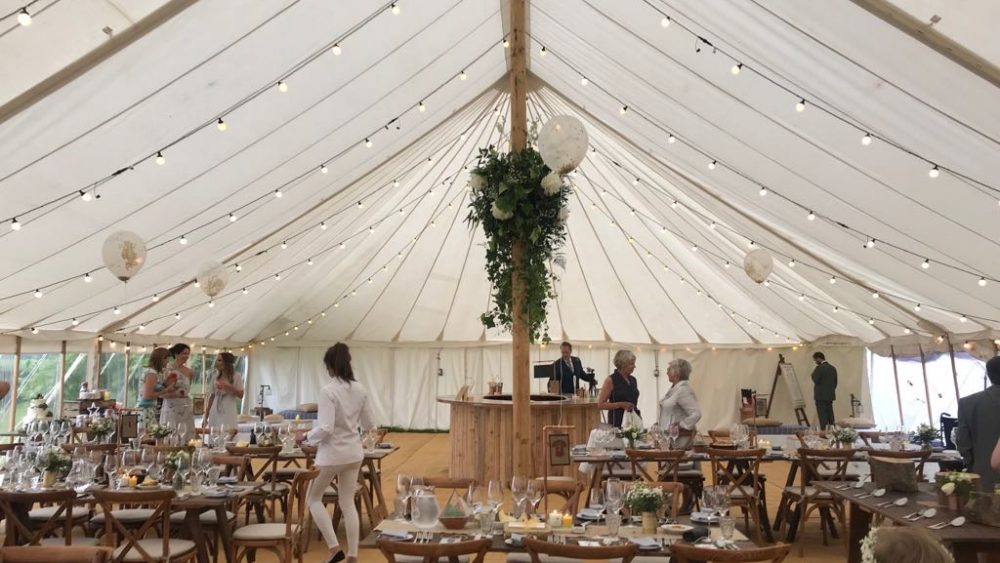 Guests filing into a traditional marquee with rustic furniture set in circles. Festoon lighting is hung around the ceiling.