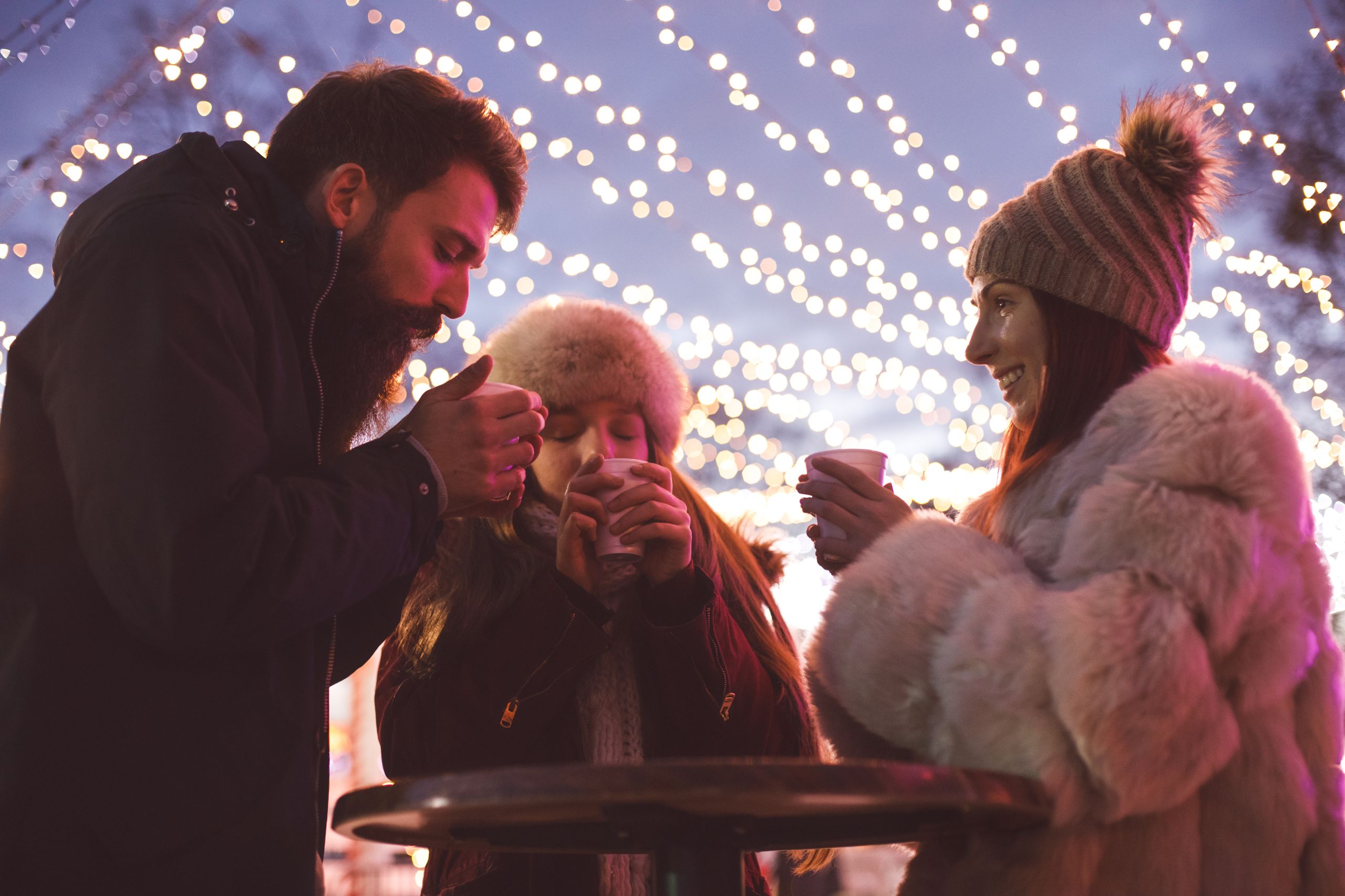 People standing around high circular table wearing winter clothing and drinking hot drinks under festoon lighting
